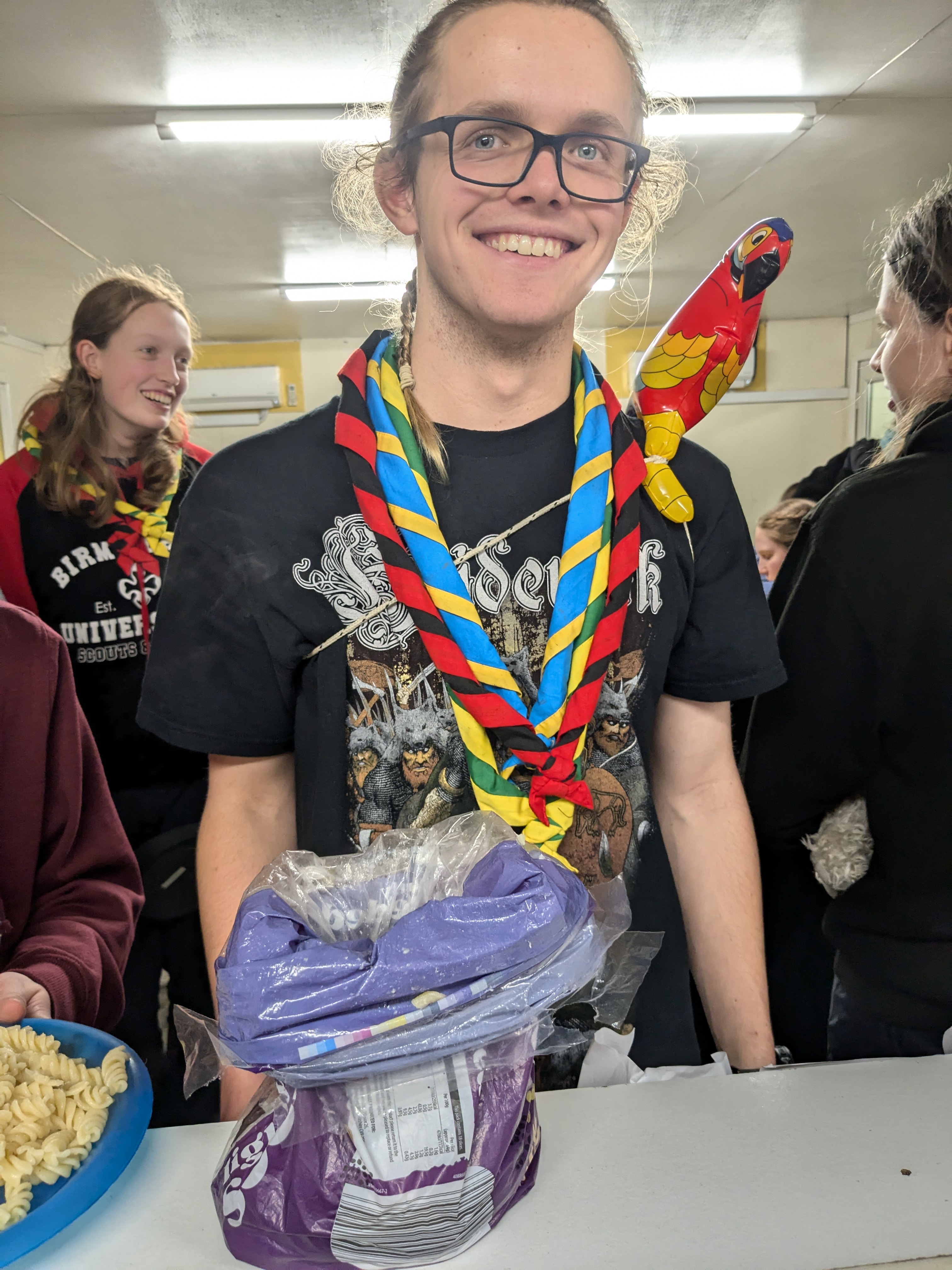Me at Midlands Freshers 2024 with my tea in a bread bag on a table, and an inflatable parrot tied to my shoulder